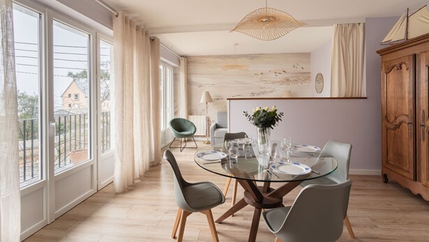 A dining area with a glass table, chairs, a vase with flowers, and a view through large windows. - Le Doux Rivage - 100 m de la plage (Dinard)