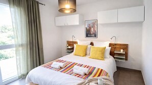 Ein modernes Hotelzimmer mit einem großen Bett, Blick auf Grünfläche, einem aufgehängten Kunstwerk und einem Nachttisch mit Lampe.