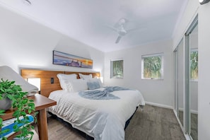 A bedroom with a bed, a bedside table with a plant, a ceiling fan, and a window with blinds. - Ocean Rise at Airlie Two (Airlie Beach)