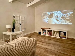 A living room with a projector screen, a white bookshelf, and a patterned ottoman.
