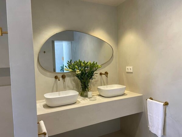 Un baño con dos lavamanos blancos tipo pedestal, un espejo ovalado grande y un pequeño florero con plantas verdes.