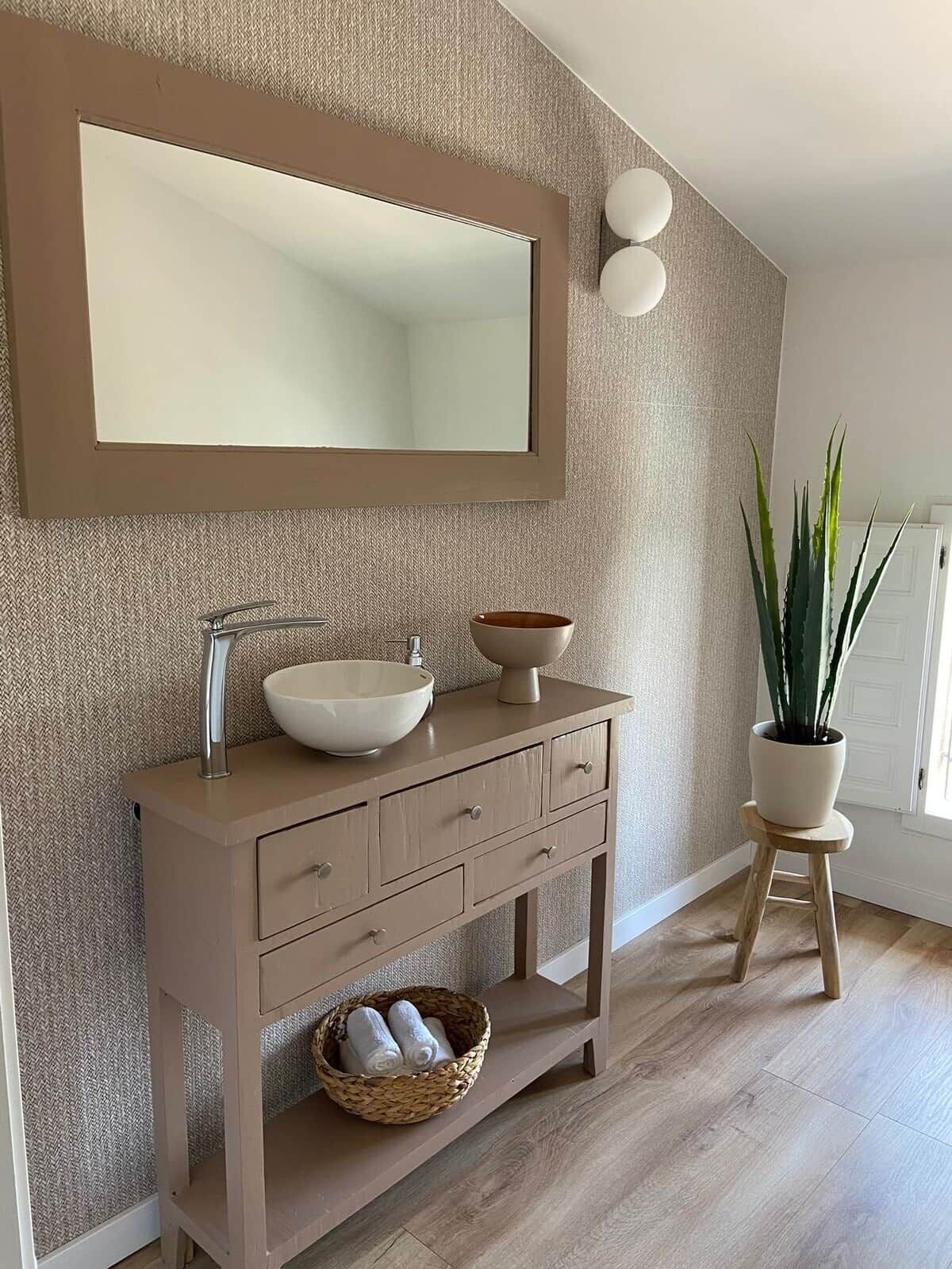 A bathroom with a beige vanity, a large mirror, a bowl sink, and a potted plant.