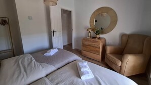 A hotel room with two beds, a wooden dresser, a round mirror, and a beige armchair.