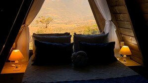 Ein gemütliches, schummriges Zimmer mit einem Bett, zwei Kissen und einem Nachttisch mit Lampe. Ein Fenster bietet Blick auf eine Berglandschaft.