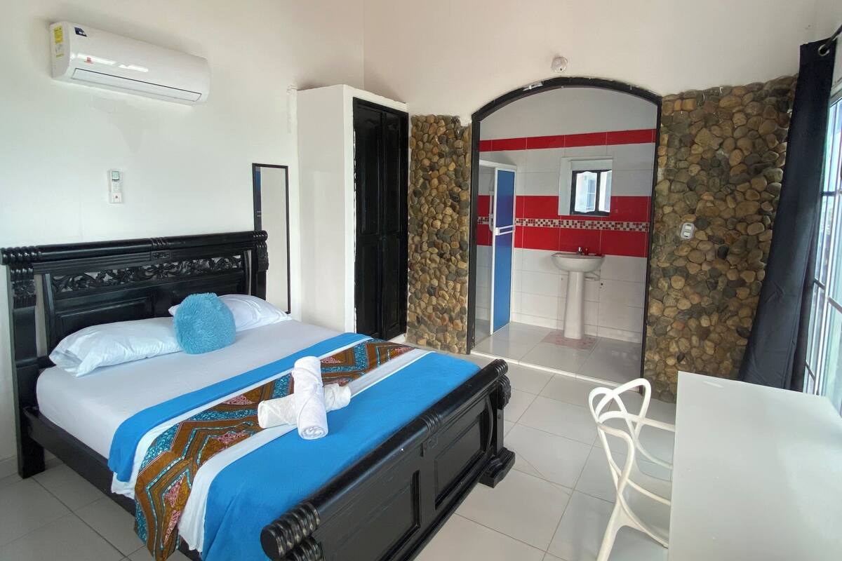 Une chambre d’hôtel comprenant un lit, un bureau, un appareil de climatisation, un mur en pierre et une salle de bain visible à travers une arche.