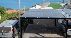 Un área de estacionamiento techada con paneles solares, un vehículo y vistas a un campo deportivo y edificios.