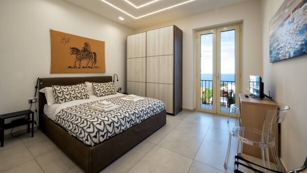A bedroom with a bed, a desk, a chair, a wardrobe, a painting, and a balcony with a view. - ZenOrlando House Apartament Sea view+Parking 2S (Taormina)