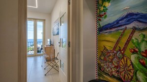 A hallway with a view to a balcony, a dining area with chairs, and a mural with a mountain and a cart. - ZenOrlando House Apartament Sea view+Parking 2S (Taormina)