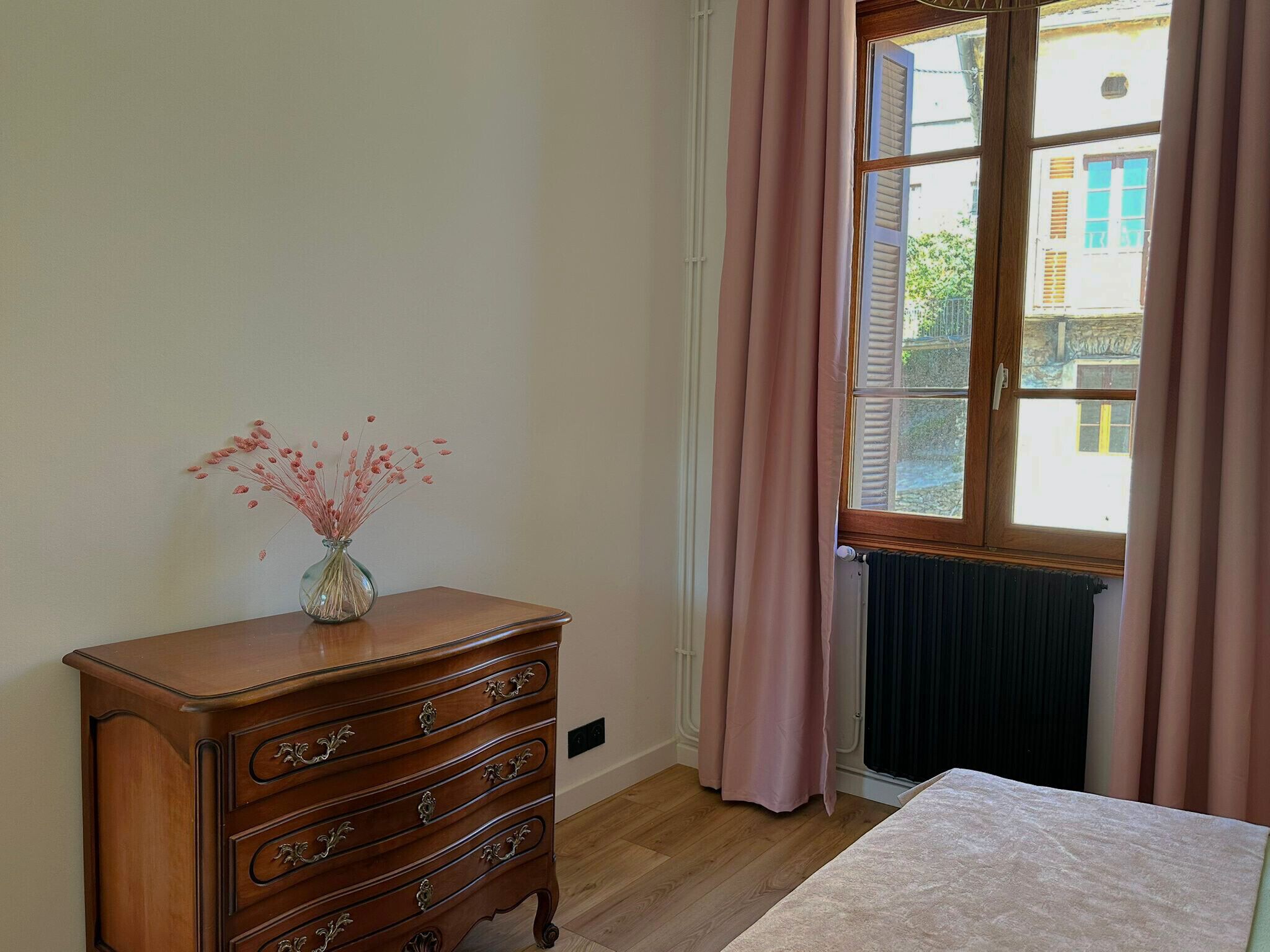 Una habitación con un aparador de madera, un florero con flores secas, una ventana con cortinas y un radiador.