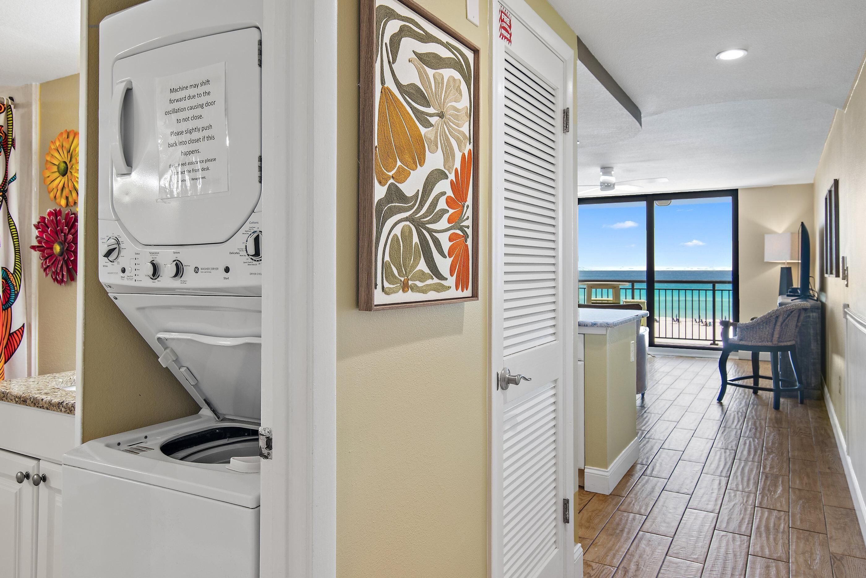 A laundry area with a front-loading washer and dryer, a door with a floral artwork, and a view of the ocean through a balcony.