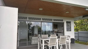 A modern outdoor patio with a glass door leading to a kitchen, white furniture, and a wooden ceiling.