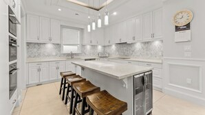 Uma cozinha moderna com armários brancos, uma ilha central com banquetas, geladeira e iluminação pendente.