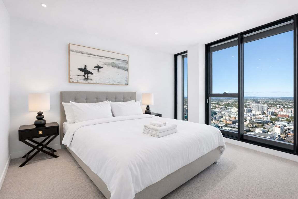 A bedroom with a large window, a bed, a nightstand, and a wall-mounted artwork.