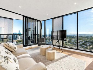 Ein modernes Wohnzimmer mit großem Fernseher, bequemen Sitzmöglichkeiten und Blick auf die Stadt durch raumhohe Fenster.