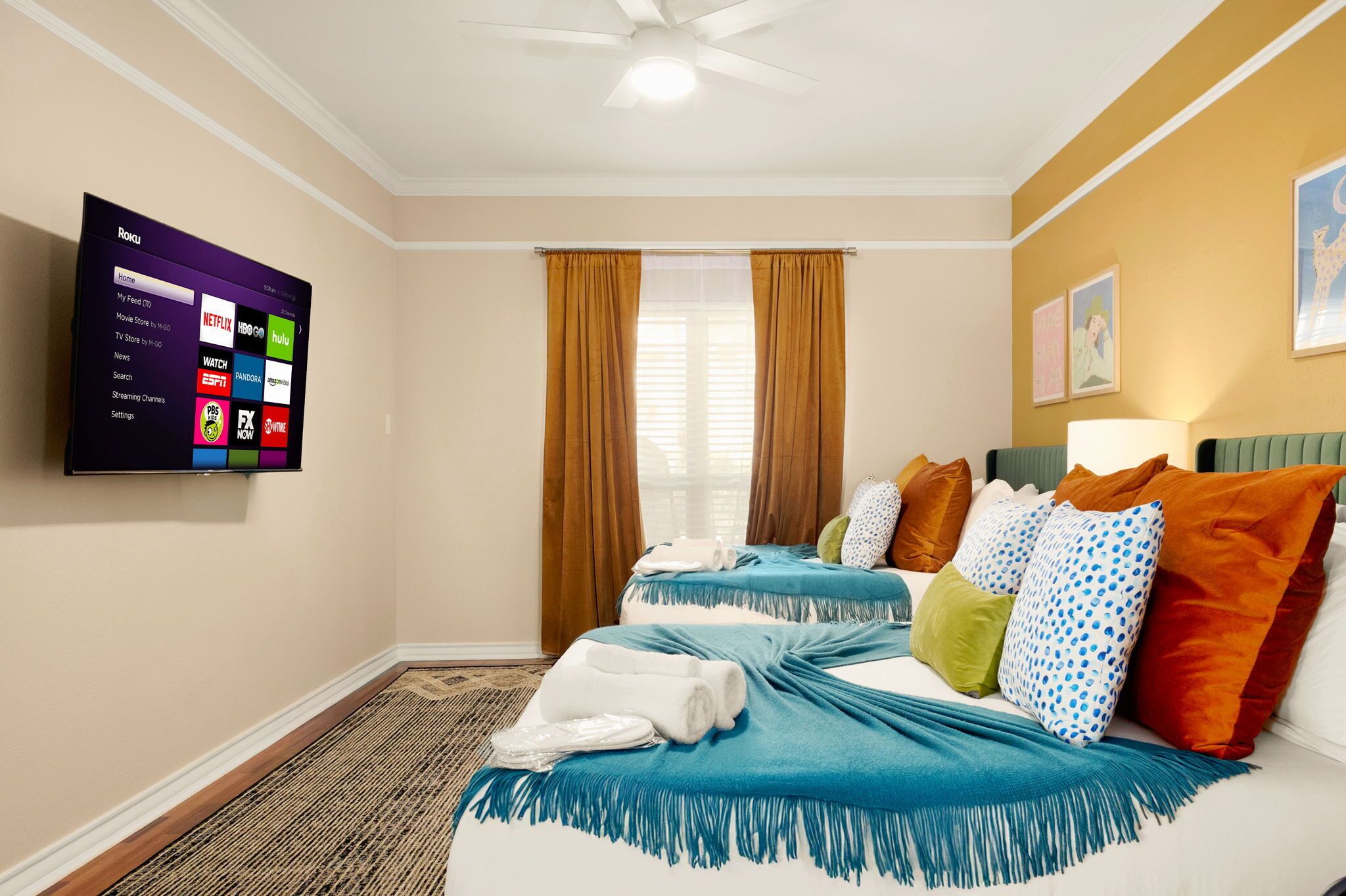 Ein Hotelzimmer mit Flachbildfernseher, der Streamingdienste anzeigt, zwei Betten mit bunter Bettwäsche, einem Fenster mit Vorhängen und einem Deckenventilator.