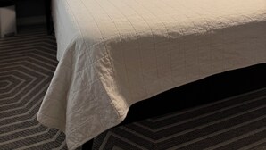 Un lit bien fait, recouvert d’une couette blanche, deux tables de chevet avec des lampes, une fenêtre avec des rideaux et une bibliothèque dans un coin.