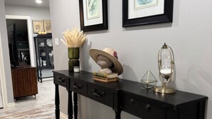 Un couloir avec une console noire, des œuvres d’art encadrées accrochées au mur, un vase contenant des plantes séchées et une tête de mannequin portant un chapeau.