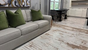 Un salon avec un canapé gris, des coussins verts, un lampadaire noir et un coin repas comprenant une table et des chaises.