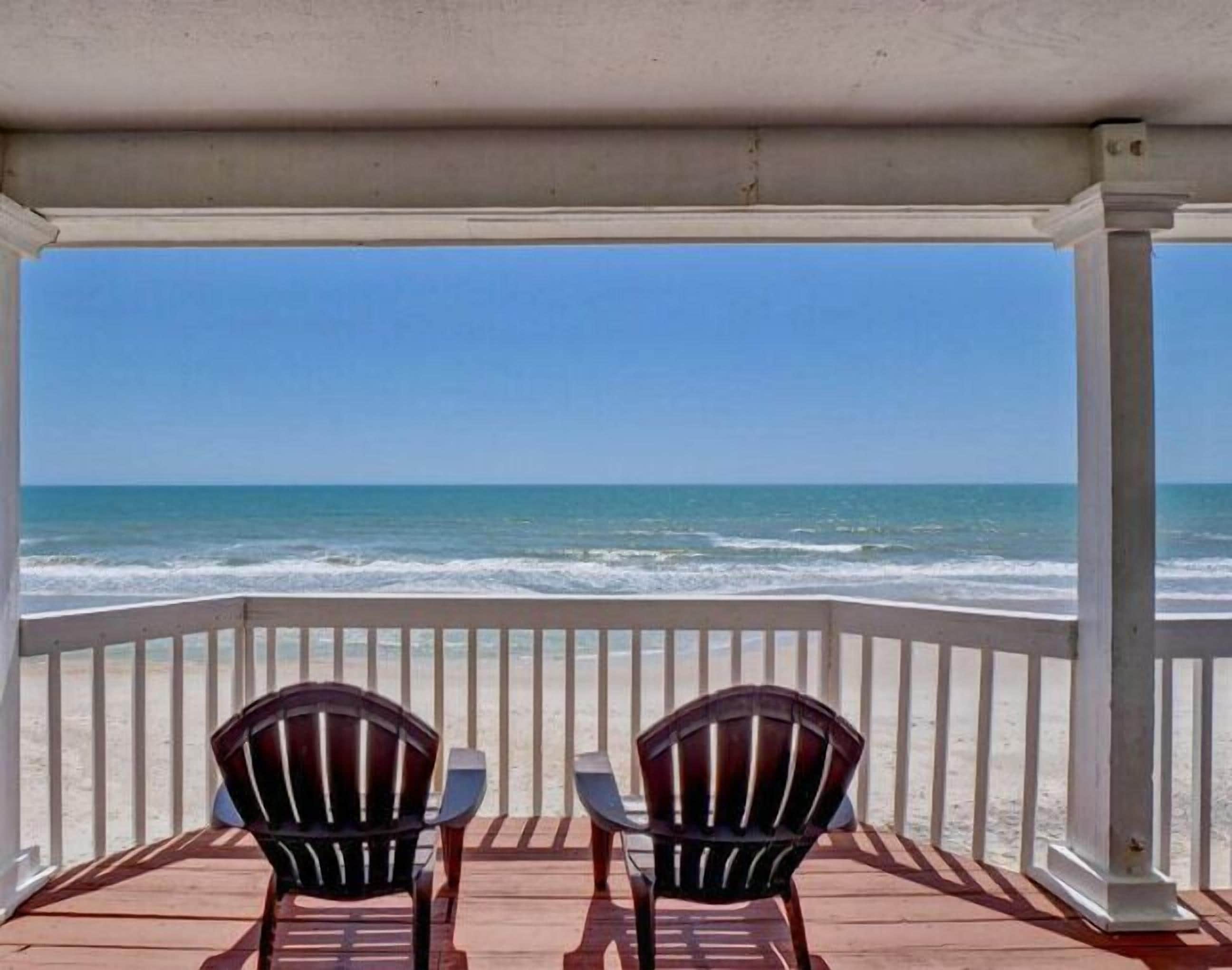 Ein überdachter Außenbereich mit zwei Adirondack-Stühlen und einem Tisch, mit Blick auf einen Strand und Wellen.