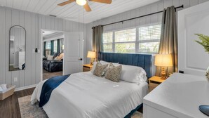 Ein Schlafzimmer mit Bett, Nachttischen, Deckenventilator, einem Fenster mit Vorhängen, einem Spiegel und einer Pflanze.