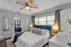 Une chambre à coucher comprenant un lit, des tables de chevet, un ventilateur de plafond, une fenêtre avec des rideaux, un miroir et une plante.