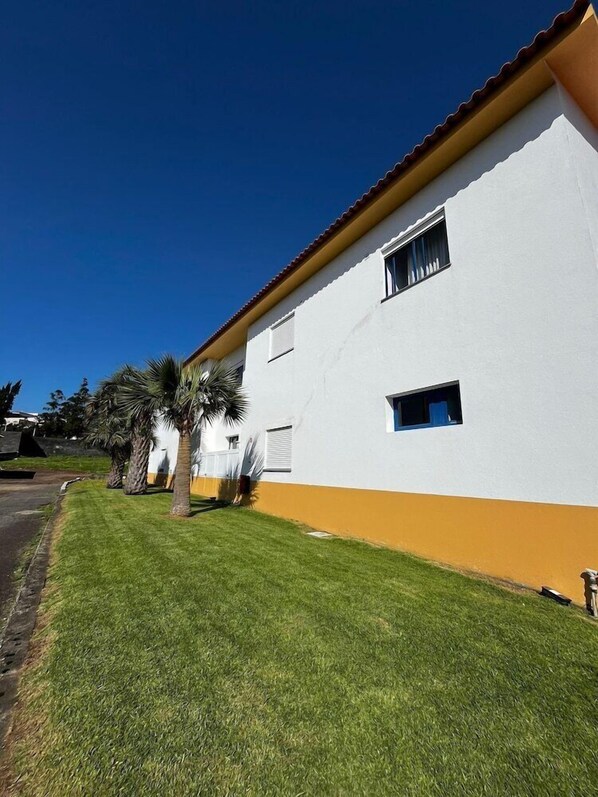 A two-story building with a white facade and orange trim, featuring a well-maintained lawn and palm trees. - Hospedaria Jsf (Horta)