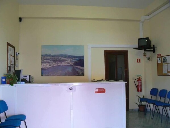 A reception area with a white counter, blue chairs, a painting of a landscape, a television, and a fire extinguisher. - Hospedaria Jsf (Horta)