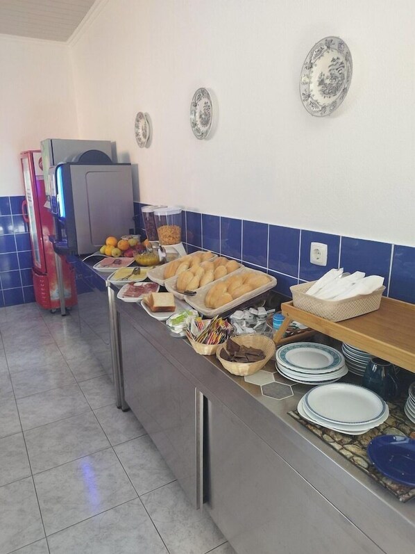 A buffet setup with various food items, a refrigerator, and stacked plates. - Hospedaria Jsf (Horta)