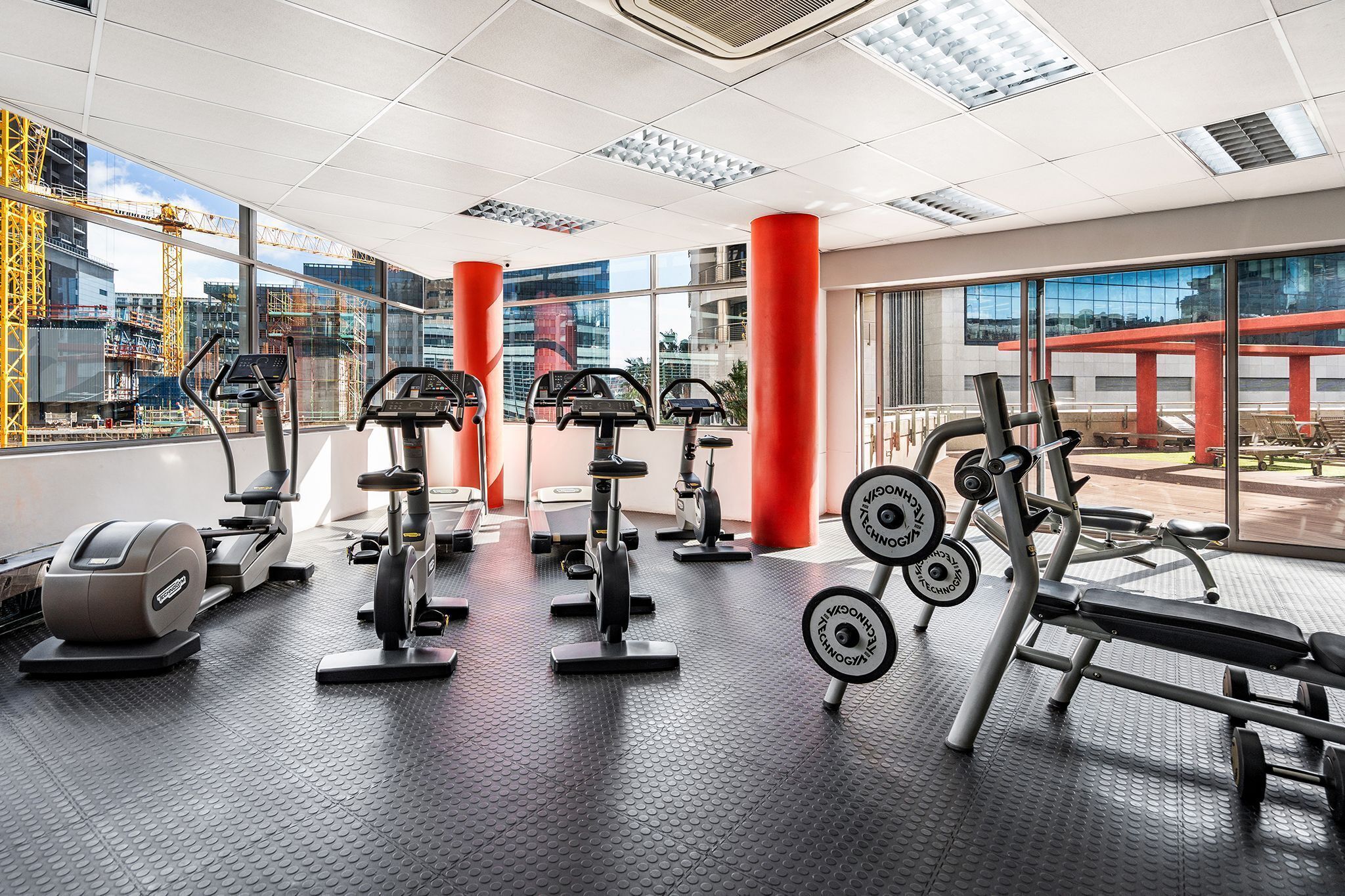 Uma academia com diversos equipamentos de exercícios, amplas janelas e vista para uma construção externa.