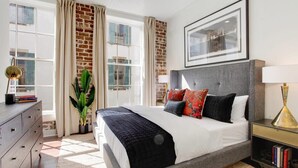 Een slaapkamer met een groot bed, een dressoir, een boekenplank, een plant in pot en een bakstenen muur.
