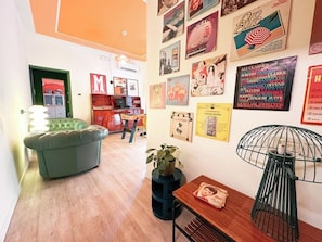 Una habitación con un sofá verde, una mesa de madera con una planta, una lámpara de metal y una pared decorada con varios carteles.