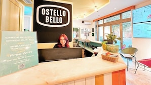 Un comptoir d’accueil avec une pancarte indiquant « OSTELLO BELLO », un tableau affichant le menu et une autre pancarte indiquant les heures d’arrivée et de départ.
