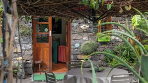 A stone-walled outdoor seating area with a table and chairs, surrounded by plants and a rustic wooden door. - Casa Belo Jardim - Studio with garden (Arco da Calheta)