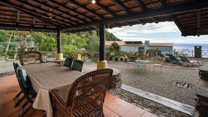 Eine Terrassengaststätte mit überdachtem Bereich, Korbstühle, einem Tisch mit Tischdecke und Blick auf Meer und Hügel.