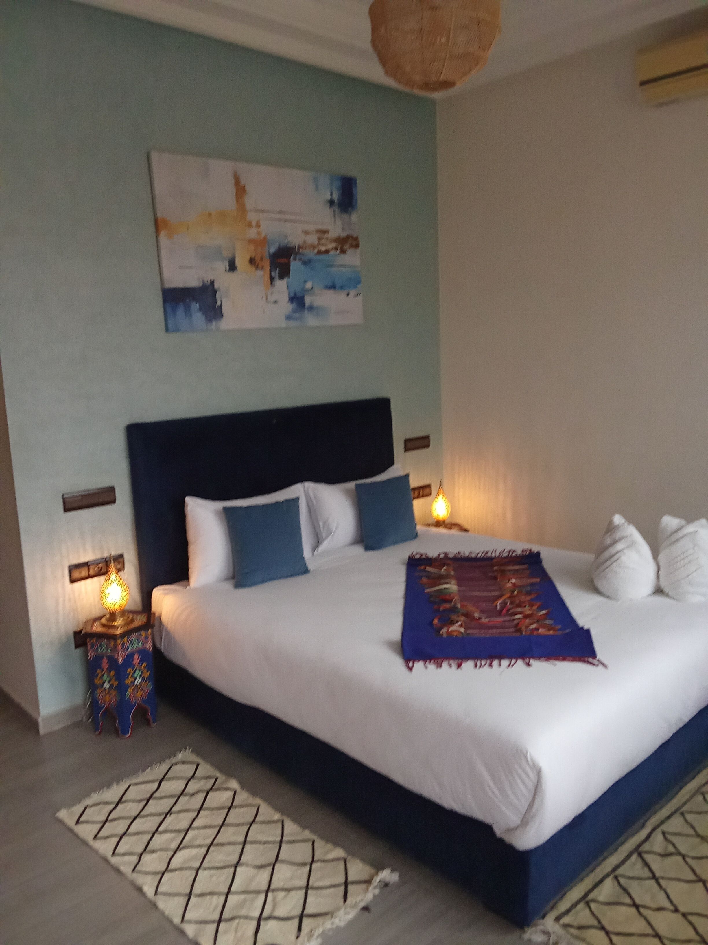 A large bed with a dark headboard, white and blue pillows, a blue throw blanket, and a wall-mounted abstract painting.