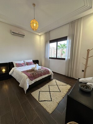 A bedroom with a bed, a bedside table, a window with curtains, a hanging light fixture, and a wall-mounted air conditioner. - Villa Amroth Marrakech: Luxury 6-Bedroom Villa with Private Pool for 14 Guests  (Marrakesh)