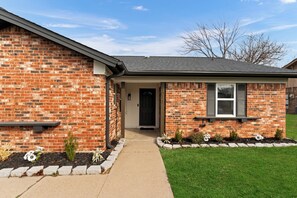 A two-story brick house with a front porch, a small garden, and a clear sky. - Bedford Bungalow Prime DFW Location Near Everything (Bedford)