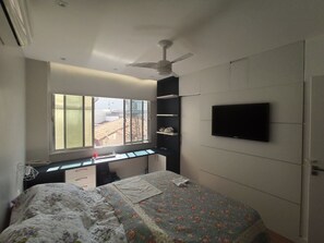 Quarto com cama, escrivaninha, cadeira, televisão, janela e ventilador de teto.