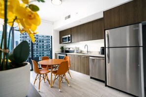 A modern kitchen with a stainless steel refrigerator, a microwave, and a sink. There is a dining table with chairs and a potted plant.
