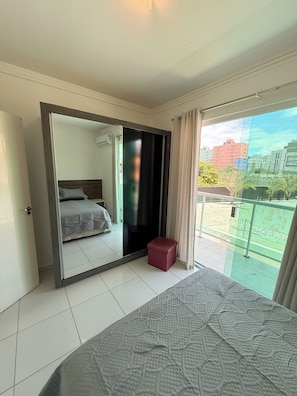 Quarto de hotel com cama, um espelho grande, porta de correr para uma varanda e um banquinho pequeno.