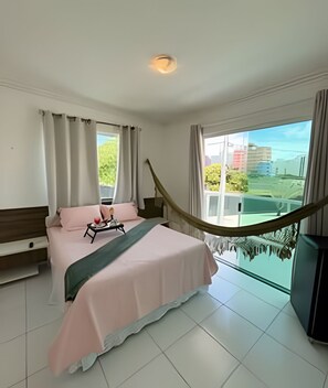 A hotel room with a bed, a small table with a tray, a TV, and a balcony with a view of buildings and greenery. - Pérola do Atlântico - Ponta de Campina (Cabedelo)