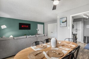A modern living room with a TV displaying 'NETFLIX,' a gray sofa, a wooden dining table with placemats, and a ceiling fan. - 0.2mi/0.3km to Dallas Stadium 2BR +FREE Parking (Arlington)