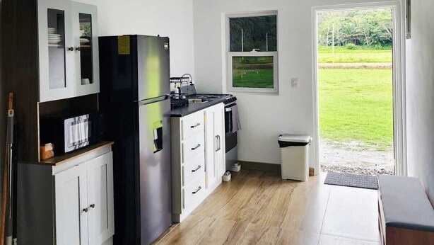 Une cuisine compacte avec des meubles blancs, un réfrigérateur noir, une cuisinière et une fenêtre donnant sur un espace verdoyant.