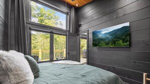 Een slaapkamer met een bed, grote ramen en uitzicht op een bos.