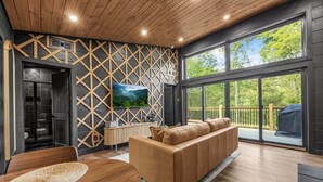 Een moderne woonkamer met een houten plafond, een grote televisie, een leren bank en uitzicht door grote ramen.