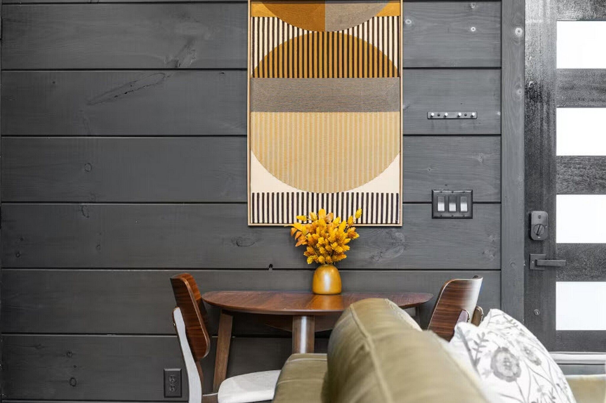 Une pièce avec un mur en bois, une table ronde en bois sur laquelle est posé un vase de fleurs jaunes, une chaise en bois et un canapé agrémenté de coussins à motifs.