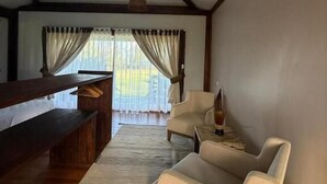 Una habitación con un banco de madera, un sofá, una mesita con una lámpara y un amplio ventanal con cortinas.