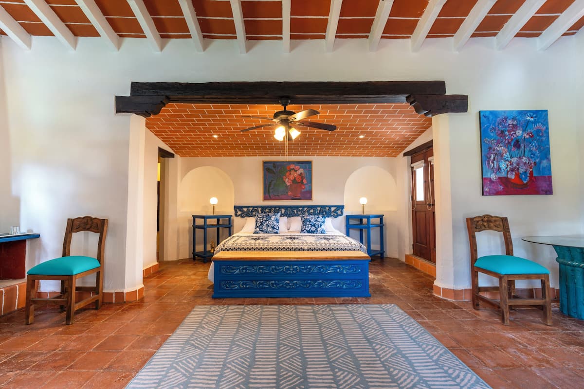A bedroom with a large bed, two chairs, a ceiling fan, and a painting on the wall.