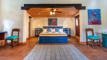 A bedroom with a large bed, two chairs, a ceiling fan, and a painting on the wall.