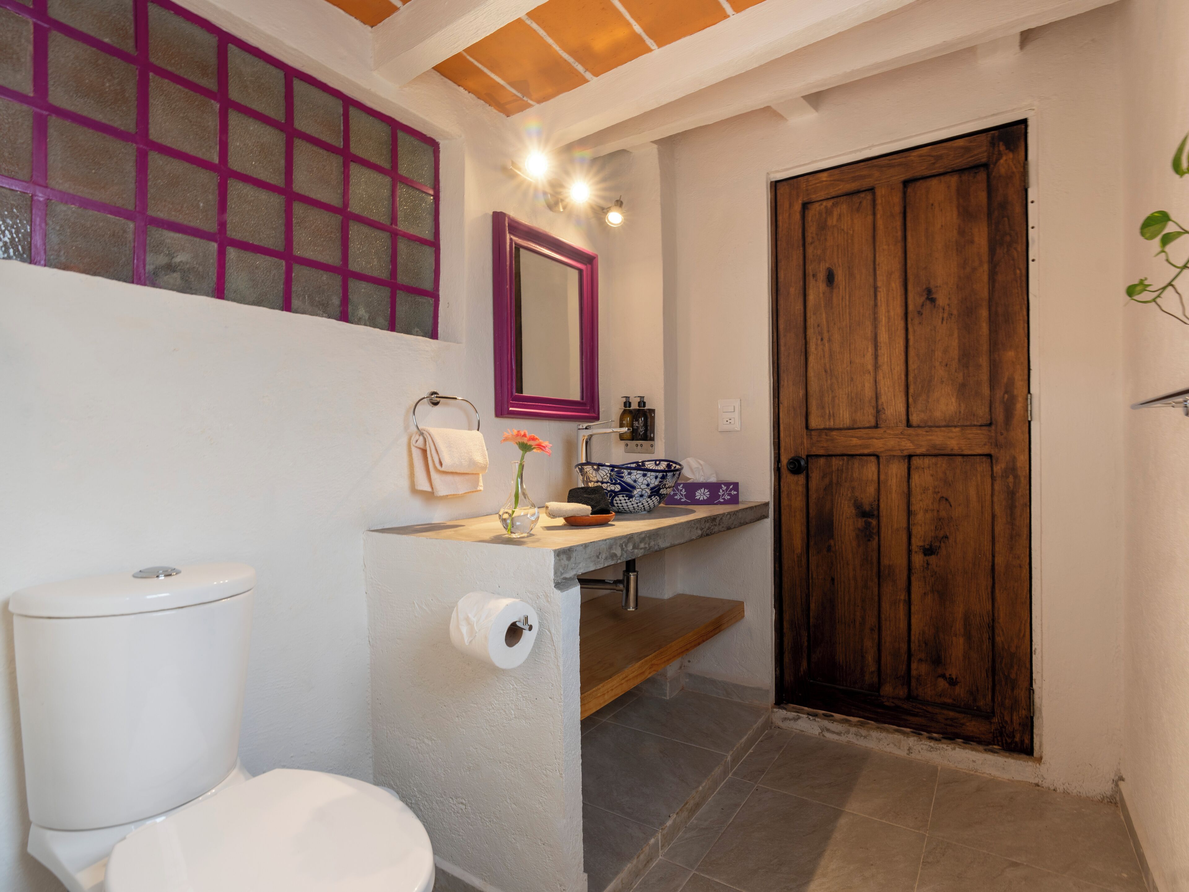 Un baño con inodoro, lavamanos con espejo, puerta de madera y ventana con diseño de celosía.